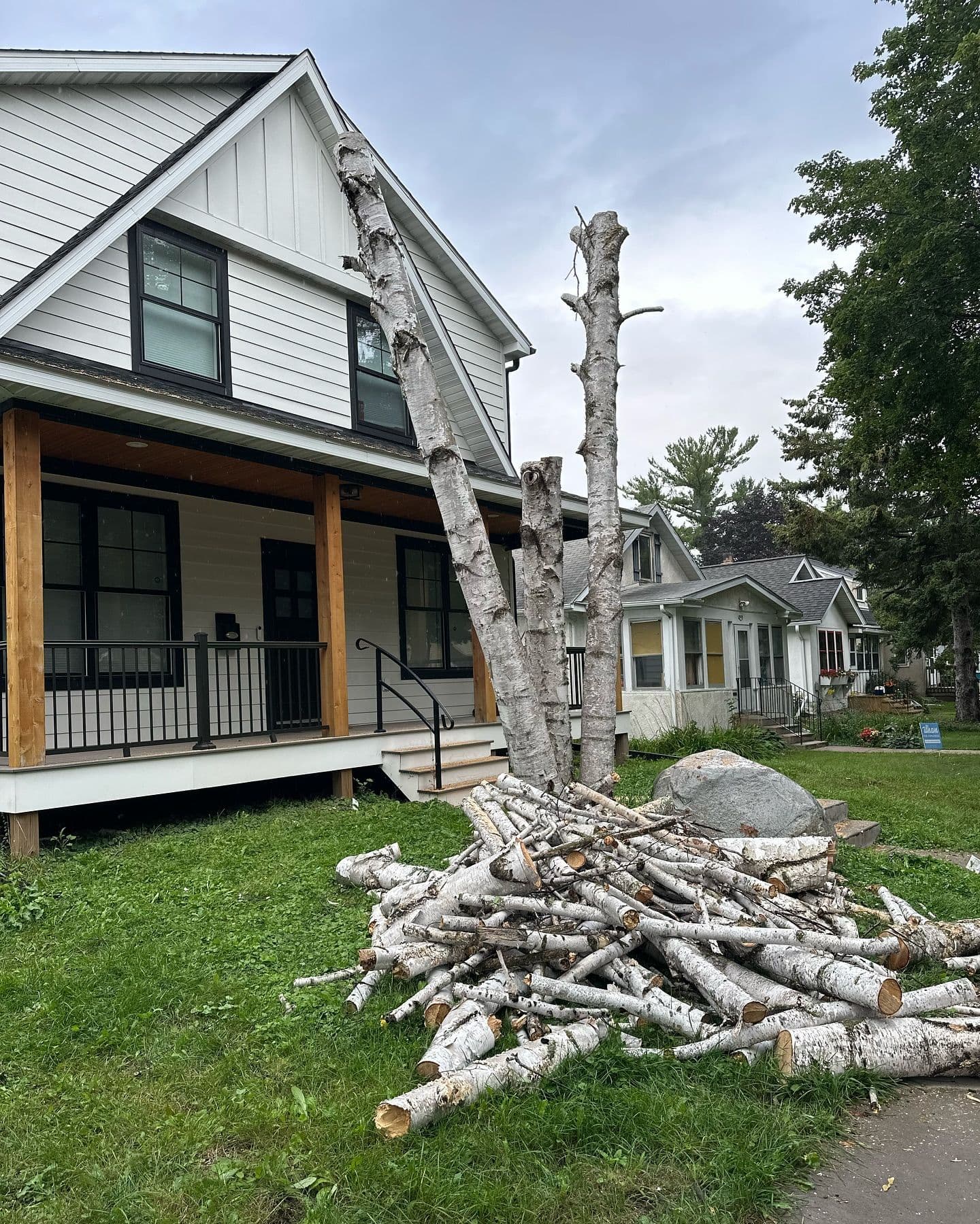 Cut birch tree logs stacked in front of a white house with a porch and green lawn.